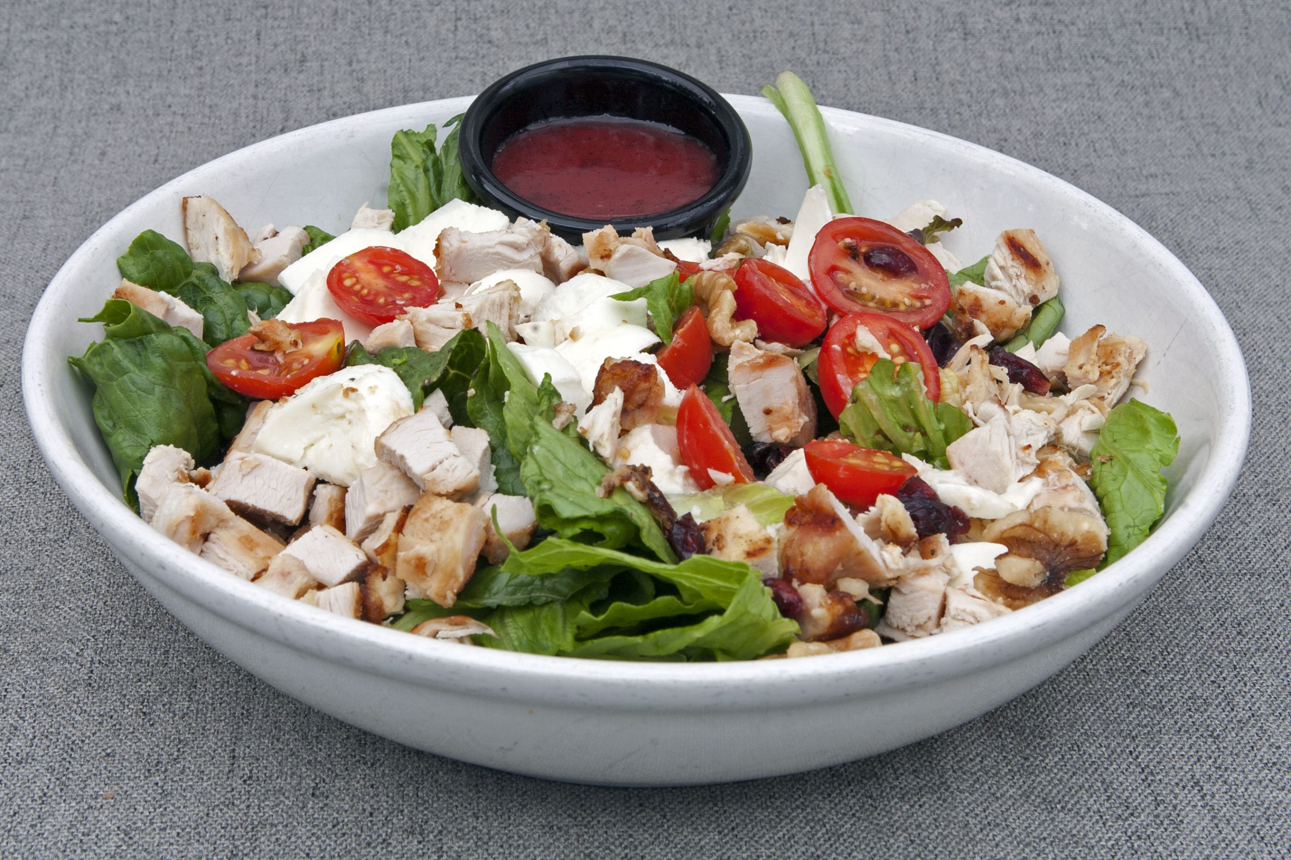 A bowl of salad with grilled chicken chunks, cherry tomatoes, cucumbers, lettuce, crumbled cheese, and a small container of dressing on a grey surface.