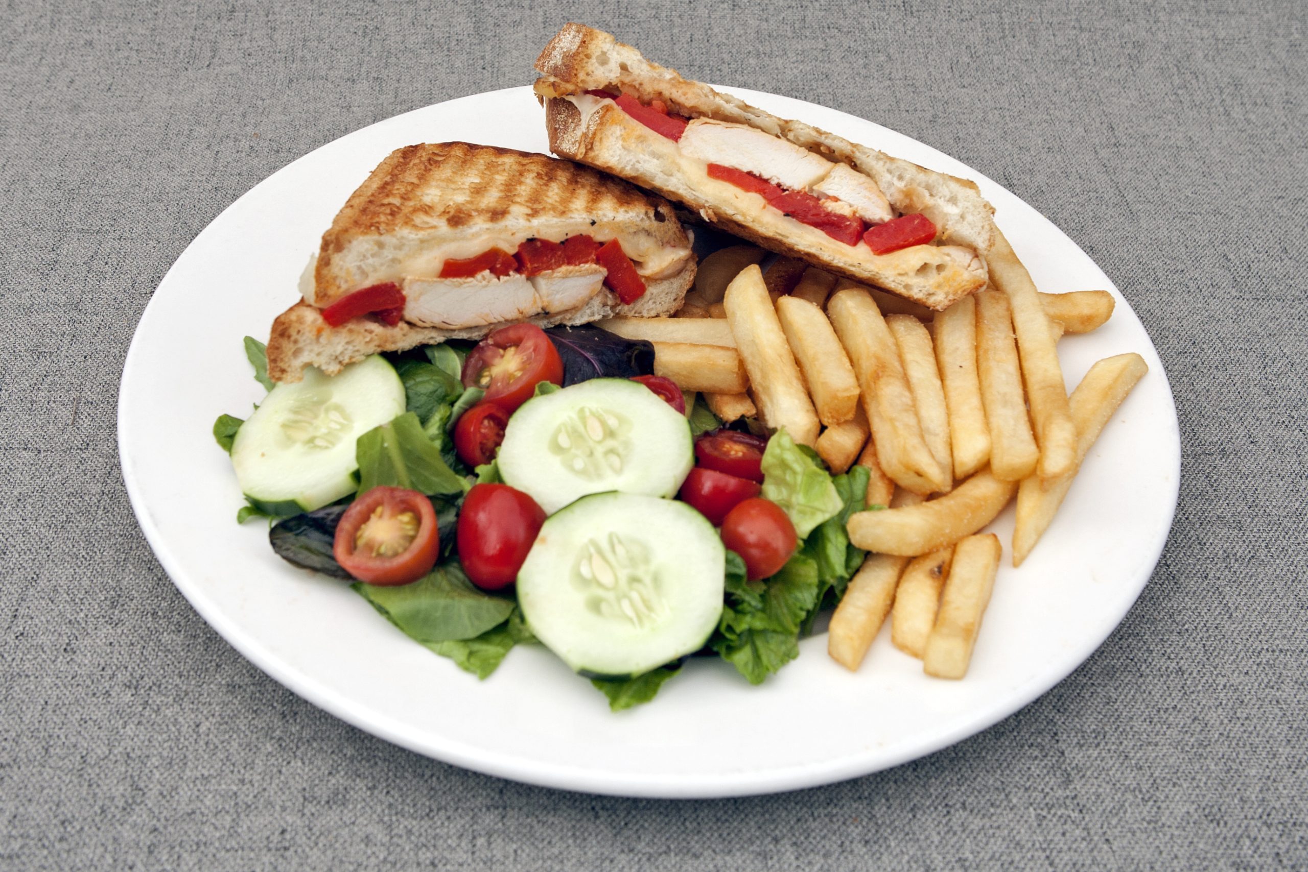 A plate with a grilled chicken sandwich, French fries, and a mixed salad containing lettuce, cucumber slices, and cherry tomatoes.